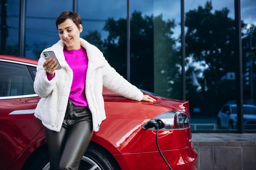 woman sitting on the hood of a red car enjoying EV charging freedom