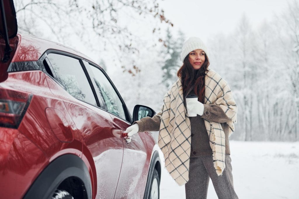 woman standing next to a red car in the snow, showcasing EV winter driving tips UK