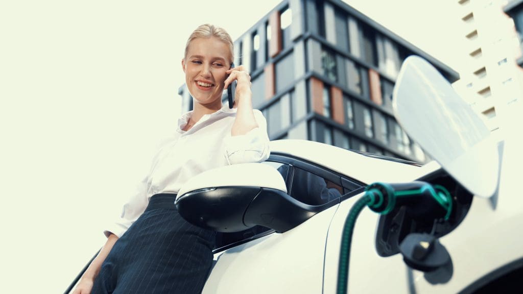 woman sitting on top of a white car talking on a cell phone in a workplace setting with ev charging in the UK