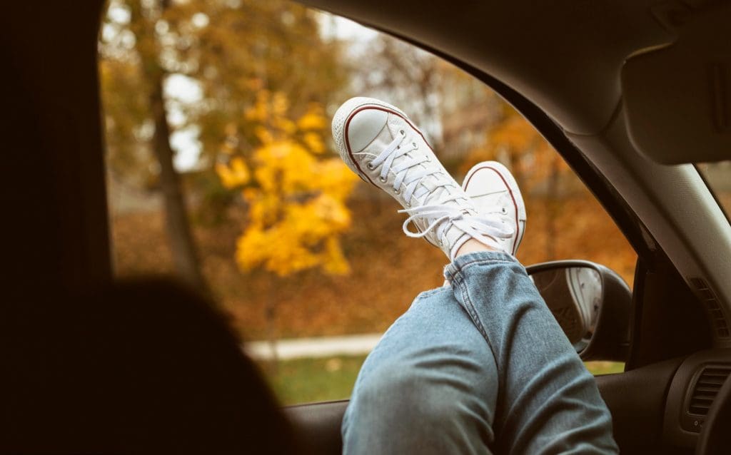 Person relaxing with their feet outside a car window beside autumn trees